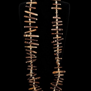May include: A long necklace featuring alternating brown, cylindrical sea urchin spines and small, off-white, spiraled seashells strung together on a thin cord. The dark brown spines vary in length and shade, creating a textured, linear pattern.