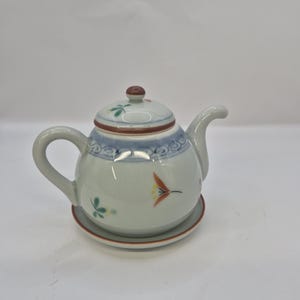 May include: A small, round, white ceramic teapot with a matching saucer. The teapot features a lid with a brown knob and is decorated with blue, red, and yellow floral designs. The teapot is designed for serving tea.