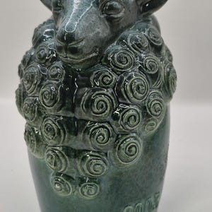May include: A ceramic sheep figurine in shades of teal and green. The sheep's face and ears are detailed, with a textured wool design. The base of the figurine has the number "2005" imprinted on it.