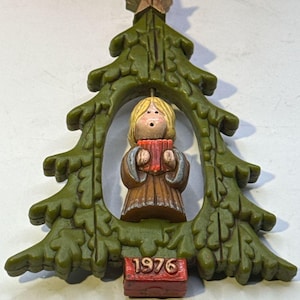 May include: A green plastic Christmas tree ornament featuring a central figure of a blonde person in a brown robe holding a red songbook. A gold star sits at the top of the tree, and a red rectangular base at the bottom displays the year 1976.