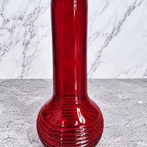 May include: A tall, cylindrical red glass vase featuring a bulbous base with horizontal ribbed detailing. The glass is translucent and smooth, with a consistent deep red hue throughout the entire structure, including the wide, open circular rim.