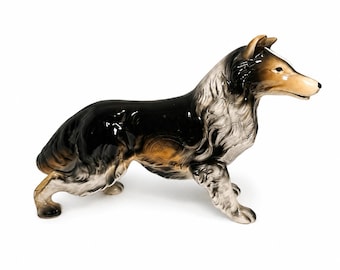 May include: A ceramic figurine of a collie dog standing in a profile position. The dog features a glossy finish with a color palette of black, white, and tan. The sculpture captures the detailed texture of the fur, pointed ears, and alert expression.
