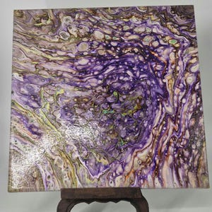 May include: Square abstract art piece with a fluid acrylic pour design. The artwork features a swirling pattern of purple, white, brown, and yellow hues. The painting is displayed on a small, dark wooden stand.