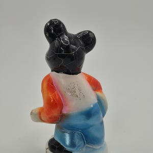 May include: A vintage figurine of a bear-like character, viewed from the back. The bear has a black head with cracked paint, an orange and white shirt, and blue pants. The figurine is on a white base.