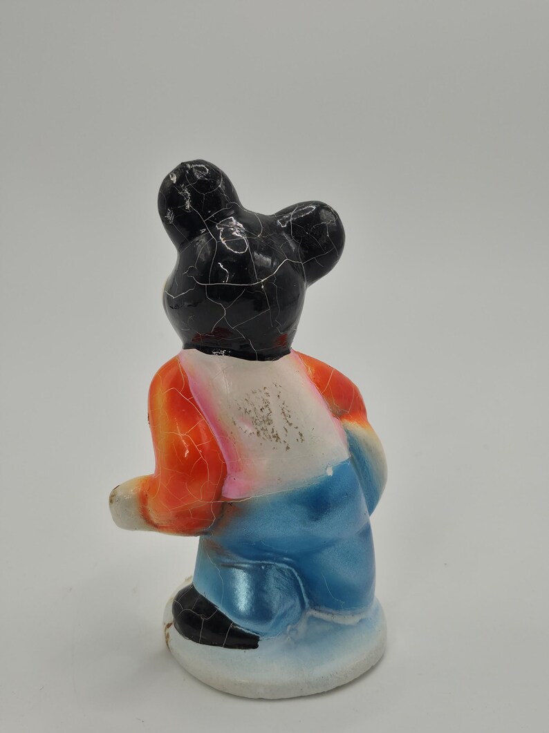 May include: A vintage figurine of a bear-like character, viewed from the back. The bear has a black head with cracked paint, an orange and white shirt, and blue pants. The figurine is on a white base.