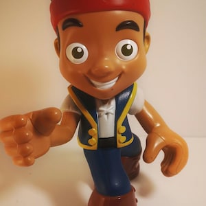 May include: A plastic figurine of a cartoon character with brown skin, black hair, and a red headband. The character wears a blue vest with yellow trim, a white shirt, and blue pants. Brown boots complete the outfit.