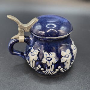 May include: A cobalt blue ceramic beer stein with a hinged metal lid and handle. The stein is decorated with white floral relief patterns. The lid is domed and the handle is curved.
