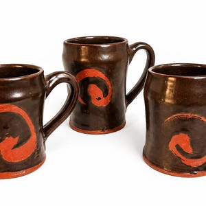 May include: Three dark brown ceramic mugs with a glossy finish, each featuring a hand-painted orange circular brushstroke design on the front. The mugs have a cylindrical body, a curved handle on the side, and a visible orange base rim.