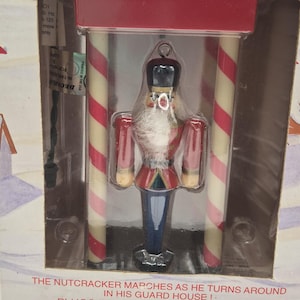 May include: A miniature animated nutcracker ornament in a clear plastic package. The nutcracker is dressed in a red jacket, blue pants, and a black hat. The ornament is set in a guard house with red and white striped columns. Text on the package reads "Animated Rotating Ornament".