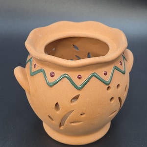 May include: A terracotta-colored ceramic pot with a wide, wavy rim and two small handles. The pot features leaf-shaped cutouts and is decorated with a green zigzag pattern and red dots. The pot sits on a small, round base.
