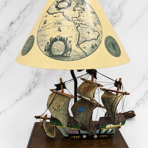 May include: A table lamp featuring a model ship base with a wooden platform and a cream-colored lampshade printed with a vintage map of the Americas, including detailed cartographic illustrations and text in a classic, historical style.
