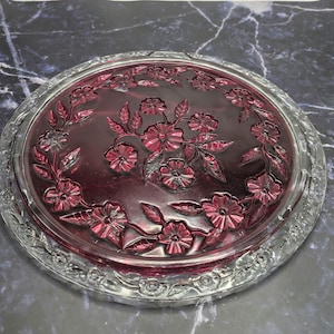 May include: A round glass plate featuring a deep purple center with an embossed floral pattern of flowers and leaves. The outer rim is made of clear glass with a decorative scalloped edge, creating a contrast between the dark center and clear border.