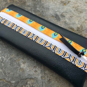 Peut inclure: Trousse rectangulaire noire avec une fermeture éclair blanche et des bandes décoratives. Une bande présente un motif géométrique gris, noir et or. L'autre bande a un motif de plumes de paon or, bleu et vert. La tirette est noire.