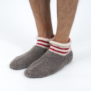 Könnte beinhalten: Graue Strick-Hausschuhe mit weißen und roten Streifen an den Bündchen. Die Socken sind aus einem weichen, bequemen Material gefertigt und perfekt, um die Füße warm zu halten.