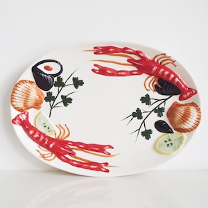 May include: An oval ceramic serving platter featuring a white background decorated with two red lobsters, two brown scallop shells, two dark blue mussels, two green parsley sprigs, and two yellow lemon slices arranged around the center.