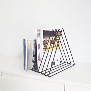 Op de afbeelding: Een zwartmetalen tijdschriftenrek in een driehoekig ontwerp, met daarin verschillende tijdschriften. Het rek is gemaakt van dunne metalen staven en staat op een wit oppervlak. De tijdschriften hebben kleurrijke covers en afbeeldingen.