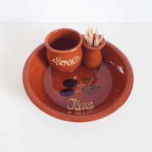 May include: A terracotta olive serving set. The set includes a round dish with a painted olive branch design and the word "Olives". Two small pots are included, one with the word "Noyaux" and the other filled with toothpicks.