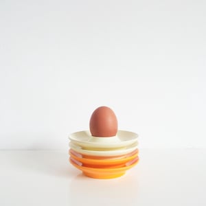 May include: A brown egg sits atop a stack of four colorful egg cups. The egg cups are in shades of cream, orange, and yellow. The egg is a light brown color. The background is white.