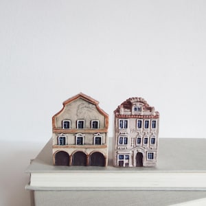 May include: Two miniature ceramic houses with detailed architectural features. One has a gabled roof and arched openings, the other a multi-story facade. Both are painted in neutral tones with brown and blue accents, resting on a stack of books.