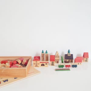 Op de afbeelding: Een houten speelgoedstad met verschillende gebouwen, waaronder huizen, torens en een kerk. De gebouwen zijn geschilderd in rood, bruin en groen en staan op een wit oppervlak. Links staat een houten kist vol met meer bouwstenen.