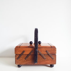 Op de afbeelding: Een rechthoekige houten naaidoos met een glanzende afwerking, voorzien van een zwart gebogen handvat en zwarte bolvormige voetjes. De doos heeft een uitschuifbaar ontwerp met metalen scharnieren aan de zijkanten en twee kleine zwarte knoppen.