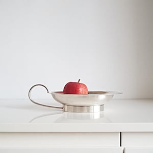 Op de afbeelding: Een glanzende zilveren serveerschaal met een handvat, met een rijpe rode appel. De schaal heeft een brede, ondiepe kom en een cirkelvormige basis. De appel is felrood van kleur, met een kleine steel.