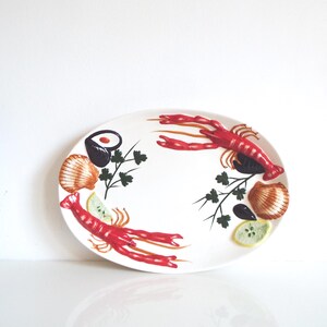 May include: An oval white ceramic serving platter featuring a colourful painted design of two red lobsters, brown seashells, dark blue mussels, green parsley sprigs, and yellow lemon slices arranged around the centre of the dish.