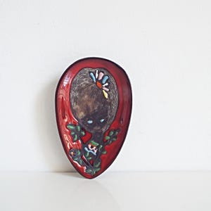 May include: A vintage, red, oval-shaped ceramic dish with a painted illustration of a girl with dark hair and blue eyes. The girl has a flower in her hair and is surrounded by green leaves and colorful flowers.
