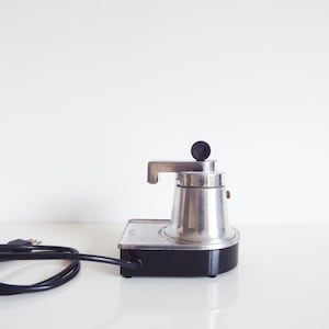 Op de afbeelding: Een elektrisch koffiezetapparaat met een zilverkleurige metalen behuizing en een zwarte voet. Het apparaat is voorzien van een zilveren tuit, een zwarte ronde knop aan de bovenzijde en een zwart netsnoer dat aan de zijkant uit het apparaat komt.