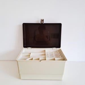 Può includere: Una scatola da cucito rettangolare con base color crema e coperchio incernierato marrone scuro. L'interno presenta un vassoio rimovibile con scomparti multipli, inclusa una sezione con perni verticali per rocchetti di filo e vari spazi.
