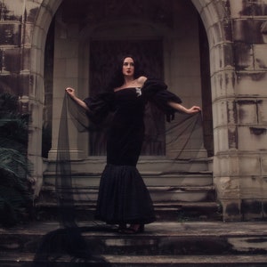 May include: A woman in a black gown with a long veil stands in front of a stone archway. The archway is part of a building with a weathered stone facade.