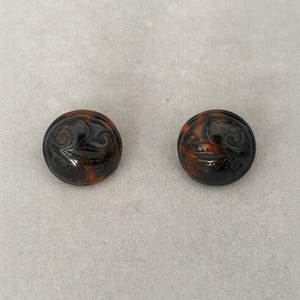 May include: Two round buttons with a tortoiseshell pattern. The buttons have a dark brown base with reddish-brown marbling and a decorative, swirling design. These buttons are suitable for sewing projects.