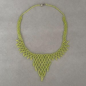 May include: A lime green beaded necklace with a delicate, openwork design. The necklace features a central, teardrop-shaped pendant and a silver-colored clasp. The beaded strands create a lacy pattern, perfect for adding a touch of elegance to any outfit.
