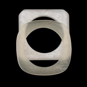 May include: Two translucent, square-shaped bangles with rounded corners, one stacked on top of the other. The top bangle is a clear, frosted material, while the bottom bangle has a marbled, off-white appearance. The background is solid black.