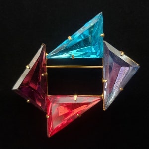 May include: A geometric brooch featuring four colorful, triangular gems in red, purple, blue, and teal, set on a gold-tone metal frame. The brooch has a pin closure on the back and is set against a black background.