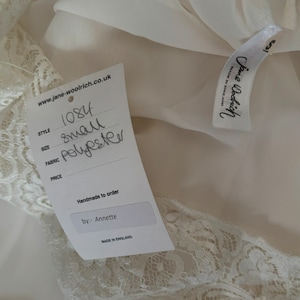Interior detail view of 2000s y2k vintage new with tags white georgette and lace sheer floor length nightgown with train by Jane Woolrich, size extra small to small. Showing the Jane Woolrich label and paper tag.