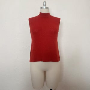 May include: A sleeveless, brick-red turtleneck sweater displayed on a mannequin. The sweater has a ribbed texture and a high neckline. The garment is a solid color and appears to be made of a soft knit material. The mannequin is white and stands against a plain white background.