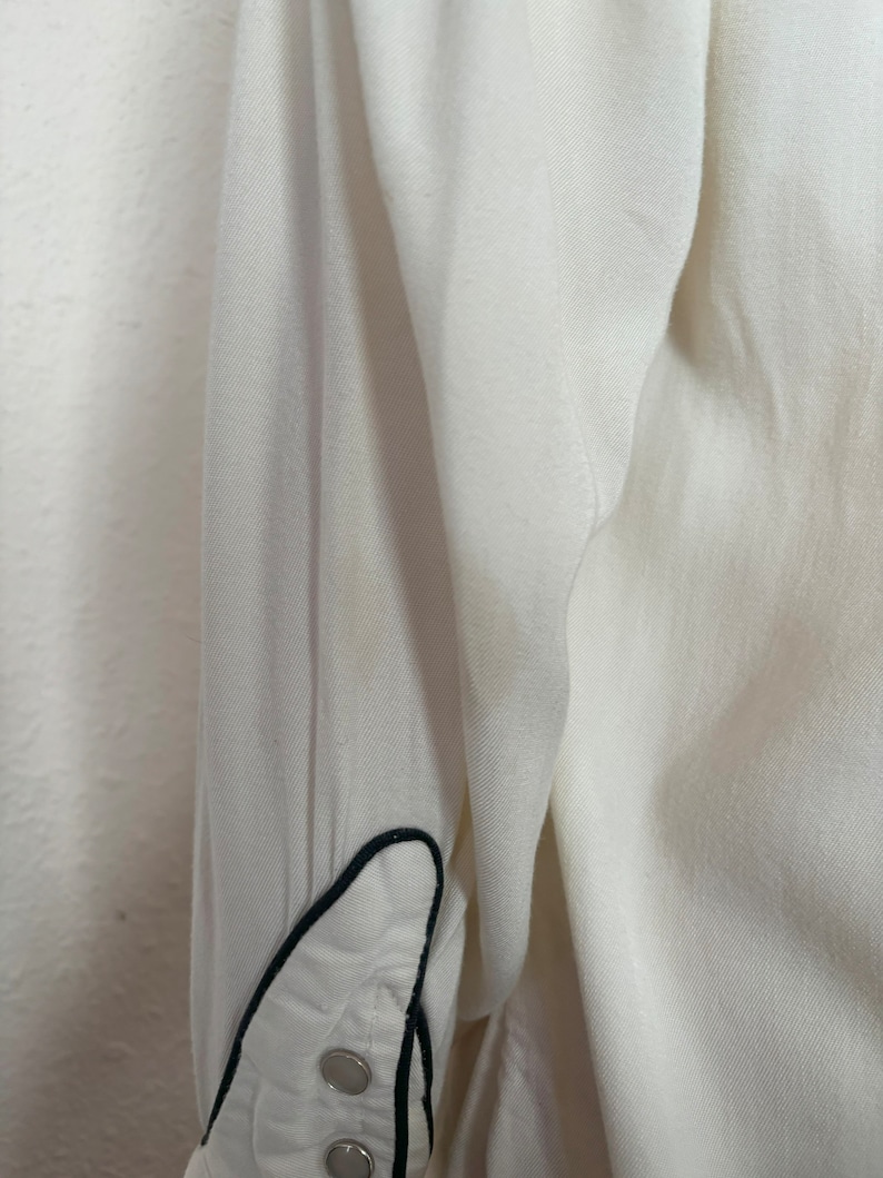 Detail view of 1960s vintage white rayon pearl snap western shirt  with navy trim and multicolored embroidery by H bar C, size small to medium. Showing a spot near the elbow.
