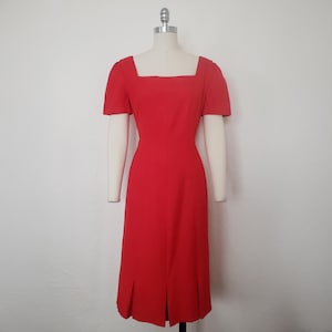 May include: A vibrant red dress with a square neckline and short sleeves. The knee-length dress features a fitted bodice and a slight A-line skirt with a slit at the back. The dress is displayed on a mannequin against a plain white background.