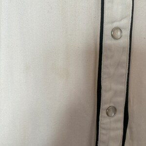 Detail view of 1960s vintage white rayon pearl snap western shirt  with navy trim and multicolored embroidery by H bar C, size small to medium. Showing a small spot on the front near the snap placket.