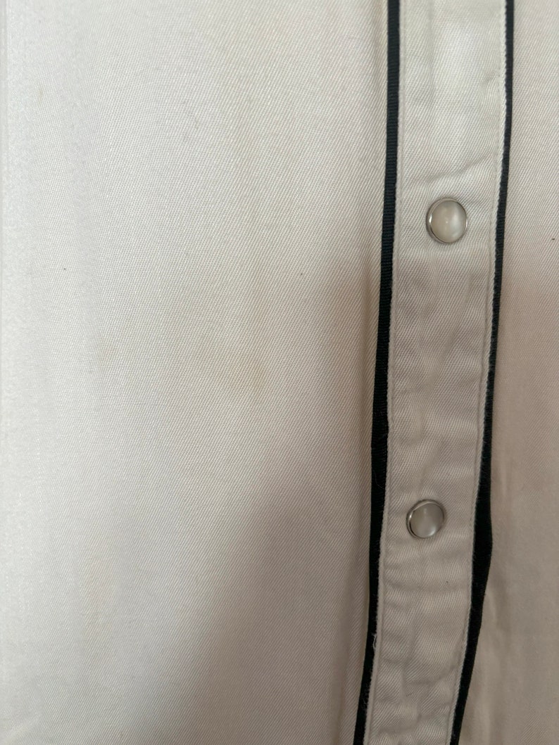 Detail view of 1960s vintage white rayon pearl snap western shirt  with navy trim and multicolored embroidery by H bar C, size small to medium. Showing a small spot on the front near the snap placket.