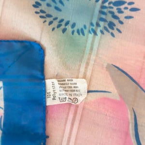 Detail view of 1980s vintage large pink, blue, and white floral print square sheer striped polyester scarf. Showing a close-up of the 100% polyester tag.