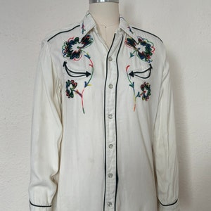 sFront view of 1960s vintage white rayon pearl snap western shirt  with navy trim and multicolored embroidery by H bar C, size small to medium.