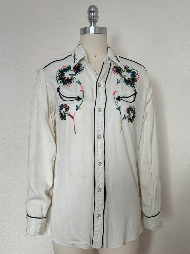 sFront view of 1960s vintage white rayon pearl snap western shirt  with navy trim and multicolored embroidery by H bar C, size small to medium.