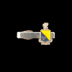 May include: Silver-toned tie clip with a shield-shaped emblem. The emblem features a yellow and blue crest with a small figure at the top and text at the bottom. The tie clip is designed to secure a tie to a shirt.