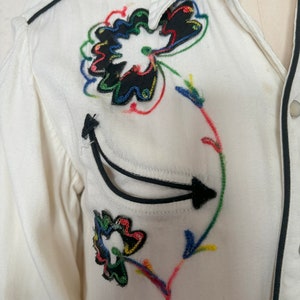 Detail view of 1960s vintage white rayon pearl snap western shirt  with navy trim and multicolored embroidery by H bar C, size small to medium. Showing the curved chest pocket and multicolor embroidery with some breaks in the thread.