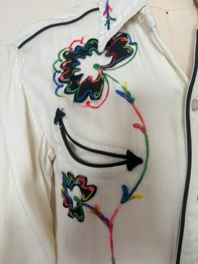 Detail view of 1960s vintage white rayon pearl snap western shirt  with navy trim and multicolored embroidery by H bar C, size small to medium. Showing the curved chest pocket and multicolor embroidery with some breaks in the thread.