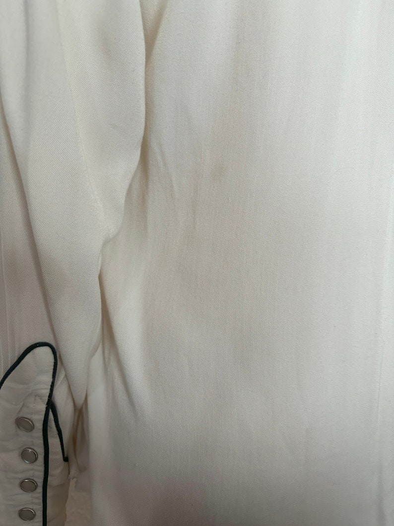 Detail view of 1960s vintage white rayon pearl snap western shirt  with navy trim and multicolored embroidery by H bar C, size small to medium. Showing a spot on the back.