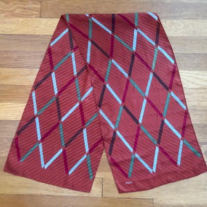 May include: A rust-colored scarf with a geometric pattern of intersecting lines in light blue, green, and dark red. The scarf is rectangular and appears to be made of a silky material. The scarf is lying on a wooden surface.