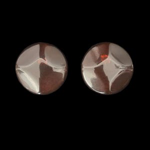 May include: Two silver-toned, round stud earrings. Each earring has a slightly domed surface with a subtle, textured design. The earrings are set against a solid black background, highlighting their reflective quality.